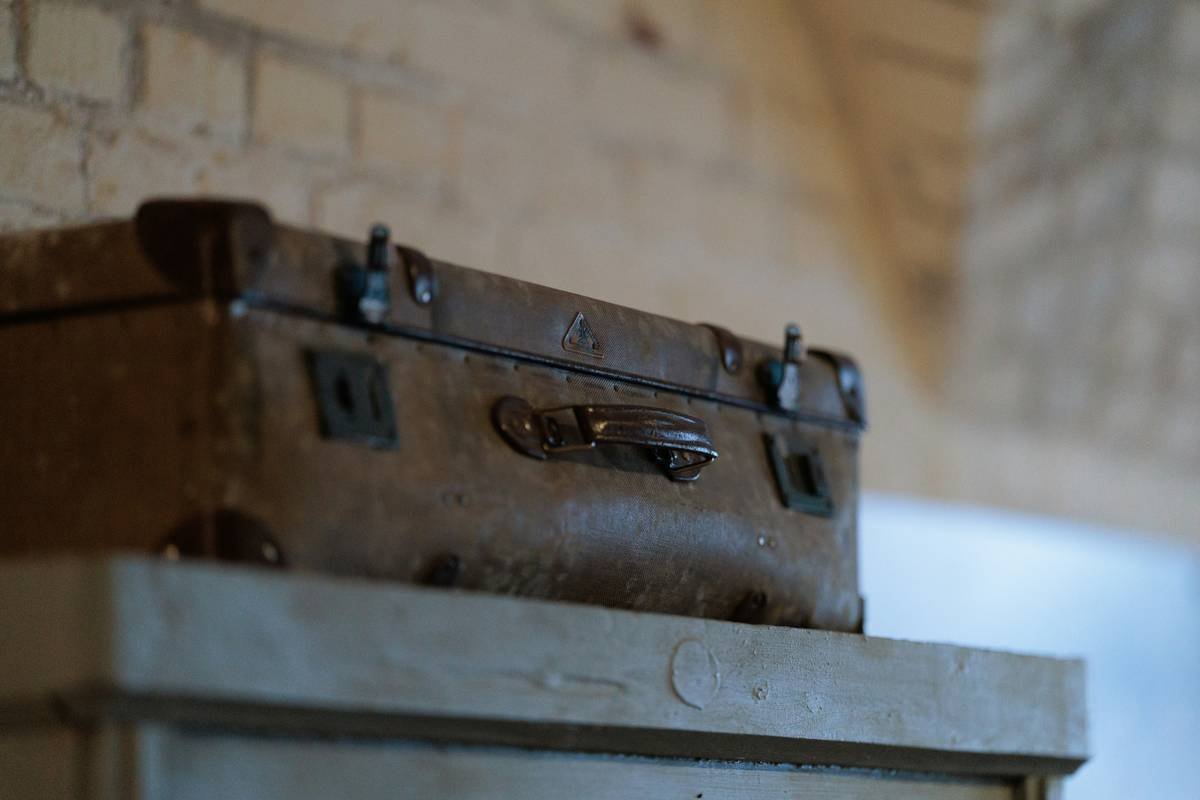 A weathered vintage tan leather suitcase rests on a shelf in front of a worn whitewashed brick wall — Konigsberg-era heritage detail.