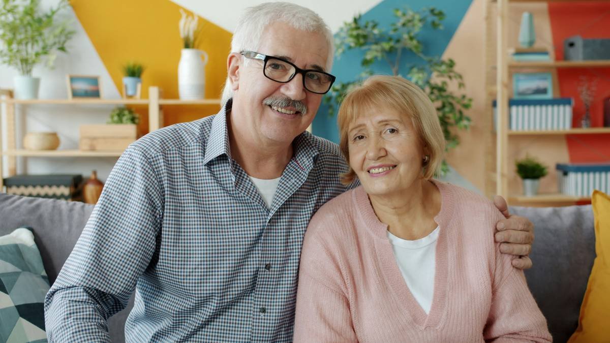 Portrait of happy retired people couple looking at camera and smiling sitting on couch at home. Committed relationship, family life and retirement concept.