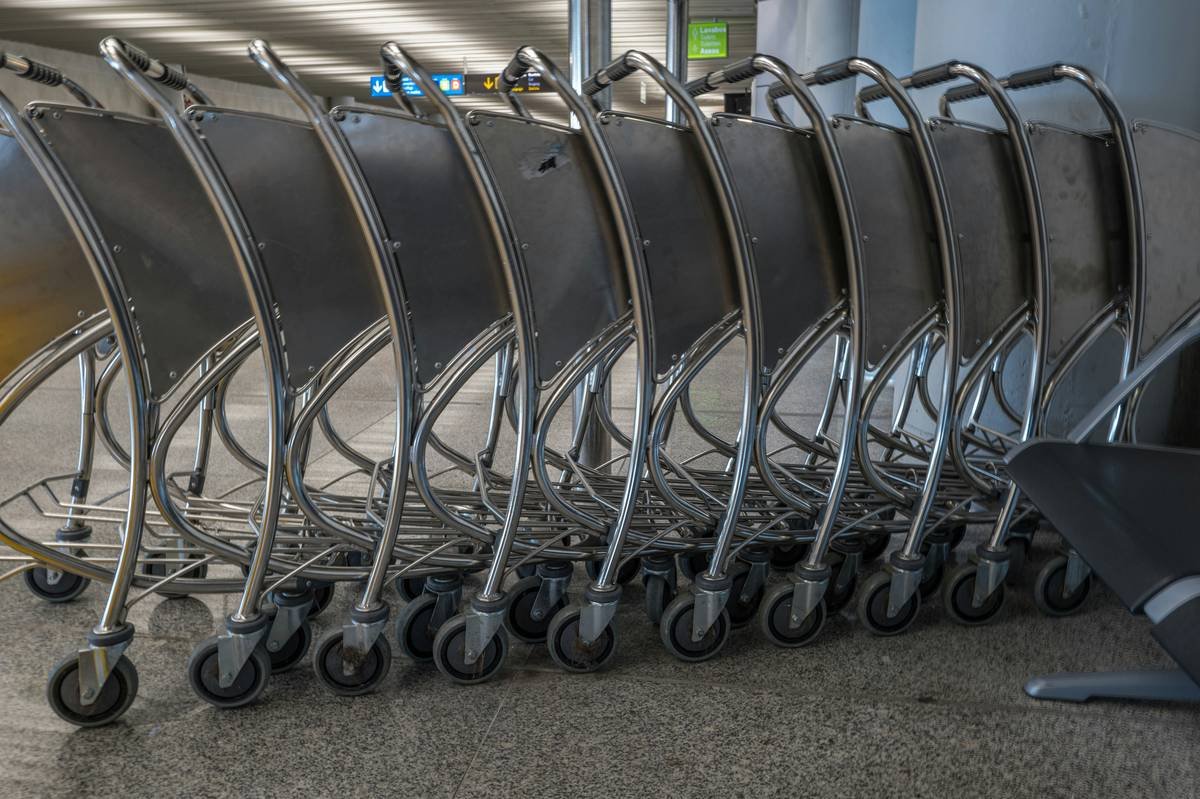 The image showcases a collection of silver airport baggage carts neatly stacked on top of each other. The carts are the primary focus, filling the frame with their metallic structure and wheels. The composition is tight, emphasizing the repetitive pattern of the carts. A sign with text in a foreign language is visible in the background, slightly blurred. The overall impression is one of order and efficiency, typical of an airport environment.