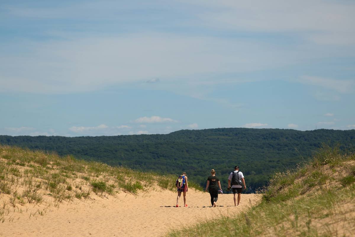 Three people walk on a sandy path between grassy dunes.